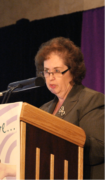 Dr-May-at-Podium-with-Purple-Background.png