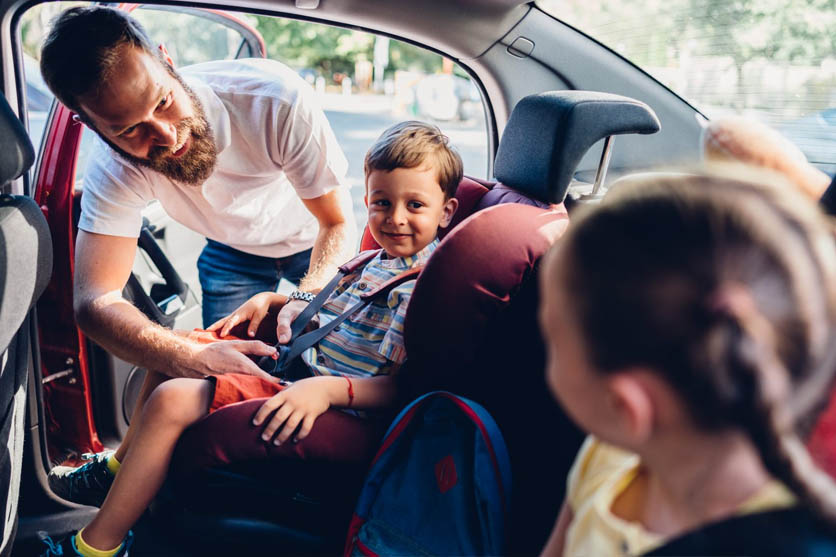 Man in white shirt buckling two kids into car seats in red car.
