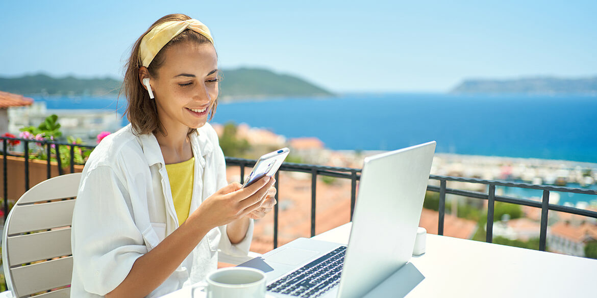 Woman in white shirt with laptop and phone wearing earbuds with ocean in background on holiday protecting from summer fraud and cyber scams.