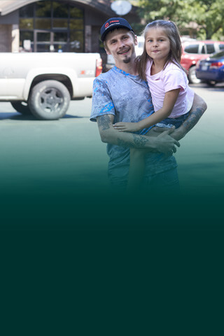 Man holding daughter shopping for truck in front of building with green swirls in background.