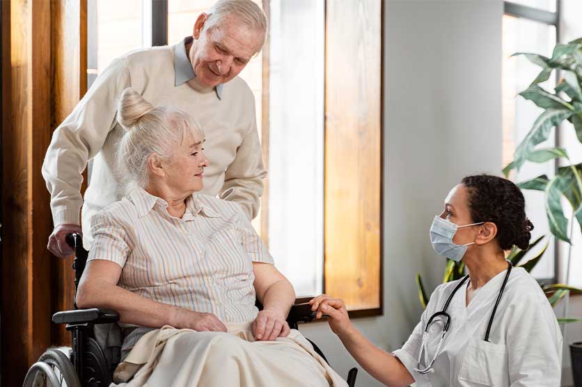 Husband behind his wife in wheelchair while long-term care nurse helps assist in care to the woman.