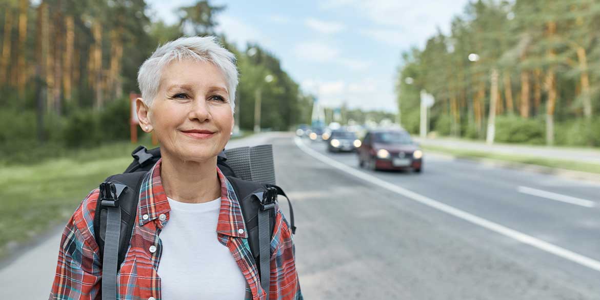 Older lady backpacking on the side of a road as she is watching for hazards on the road during her retirement