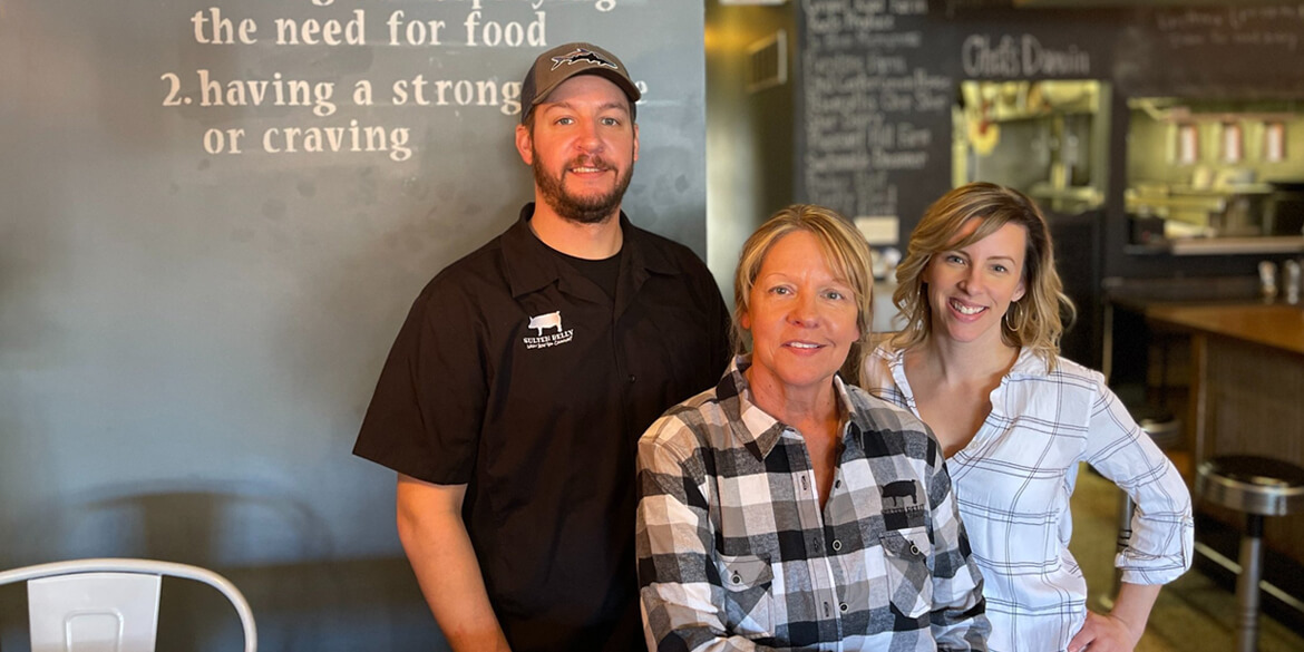 Man in black shirt and hat with two women in checked shirts in front of chalkboard menu at Sulten Belly restaurant in Waupaca Wisconsin.