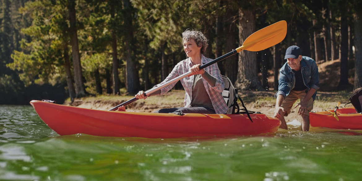 Retired woman in red kayak with paddle and man standing behind.