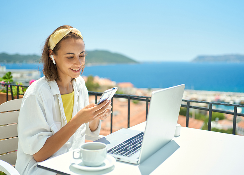 Woman in white shirt with laptop and phone wearing earbuds with ocean in background protecting from cyber fraud and cyber scams.