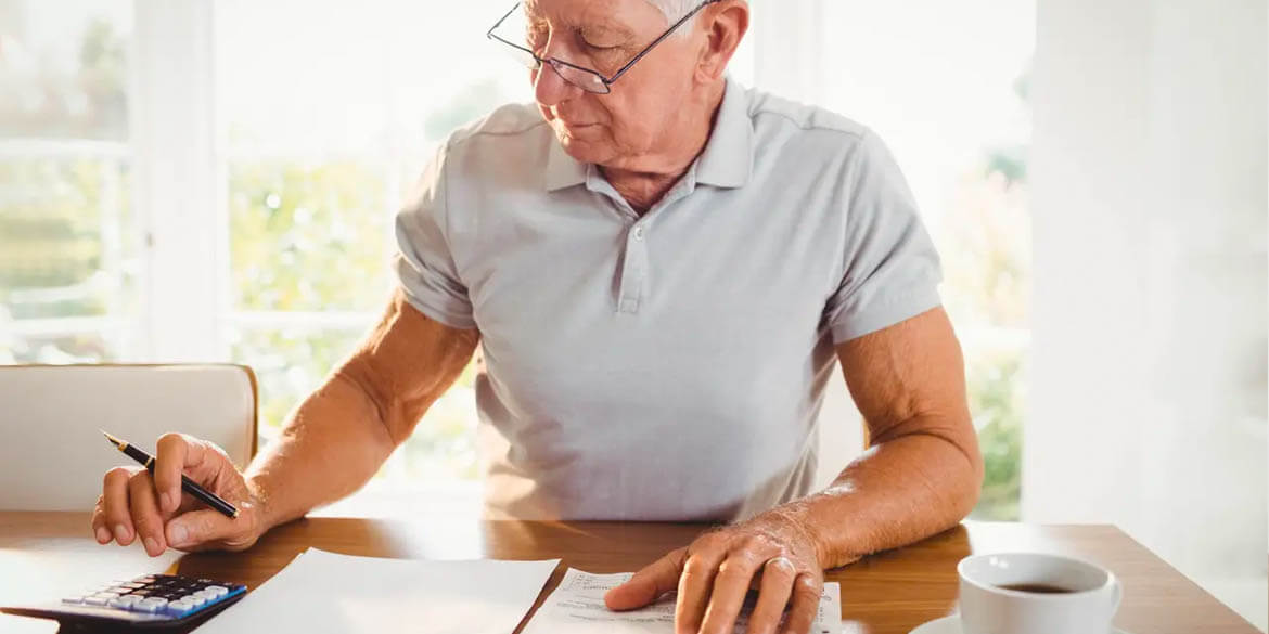 Man in white polo shirt with glasses at wood table with calculator, papers, pen and white cup of coffee.