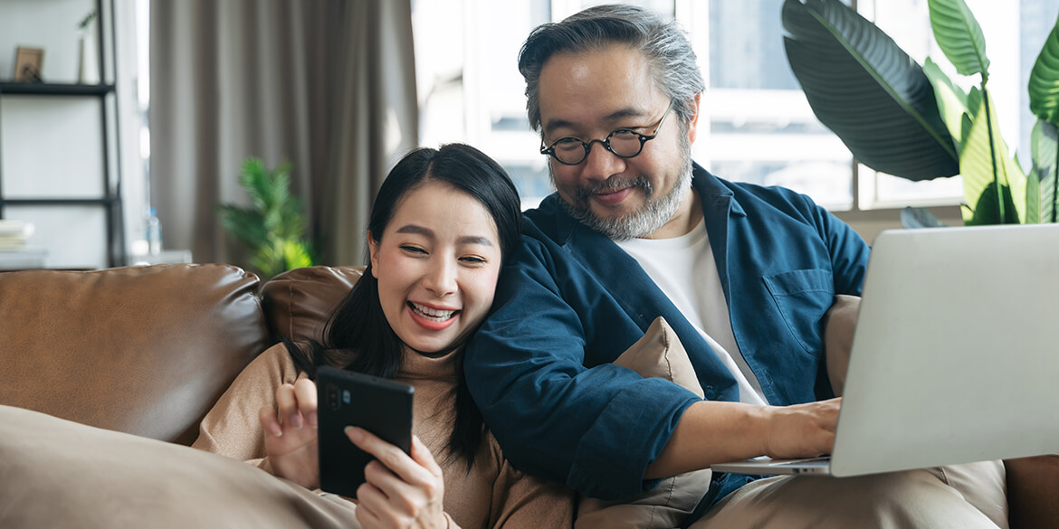 Man with glasses and blue shirt on laptop with woman in pink shirt on phone sitting on brown couch.