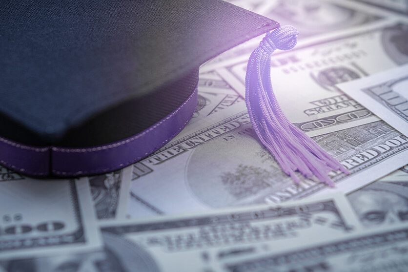 Black graduation cap with purple tassel on top of $100 bills.