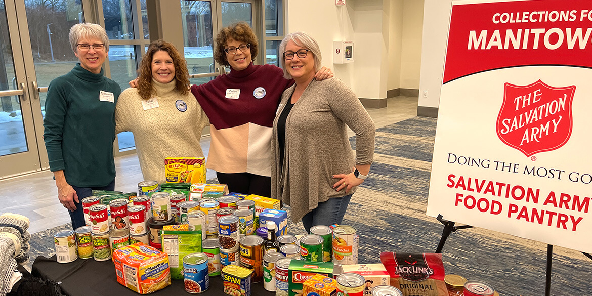 Four females in front of table full of canned and box food next to red Salvation Army poster.