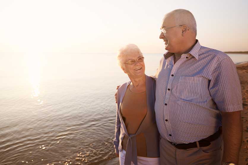 Older couple arm in arm walking on the beach with water behind them to show enjoying their social security benefits 