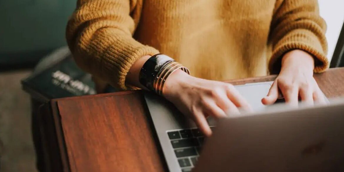 Hands with bracelets typing on laptop wearing gold sweater.