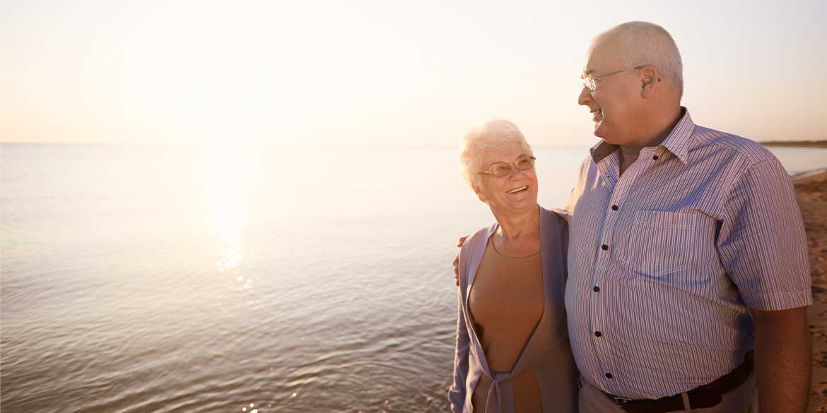 Older couple arm in arm walking on the beach with water behind them to show enjoying their social security benefits 