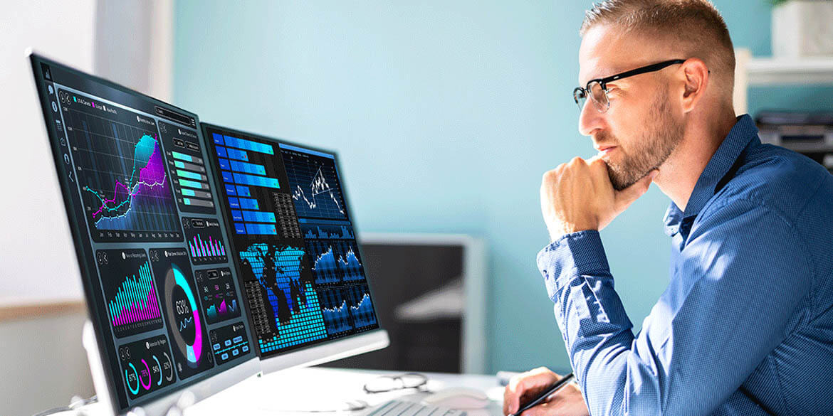Man in blue shirt and glasses looking at monitors with charts and graphs.