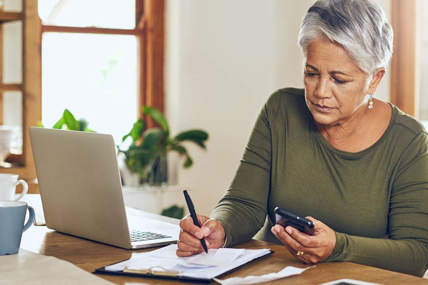 Woman with green shirt holding phone and pen near laptop and coffee.