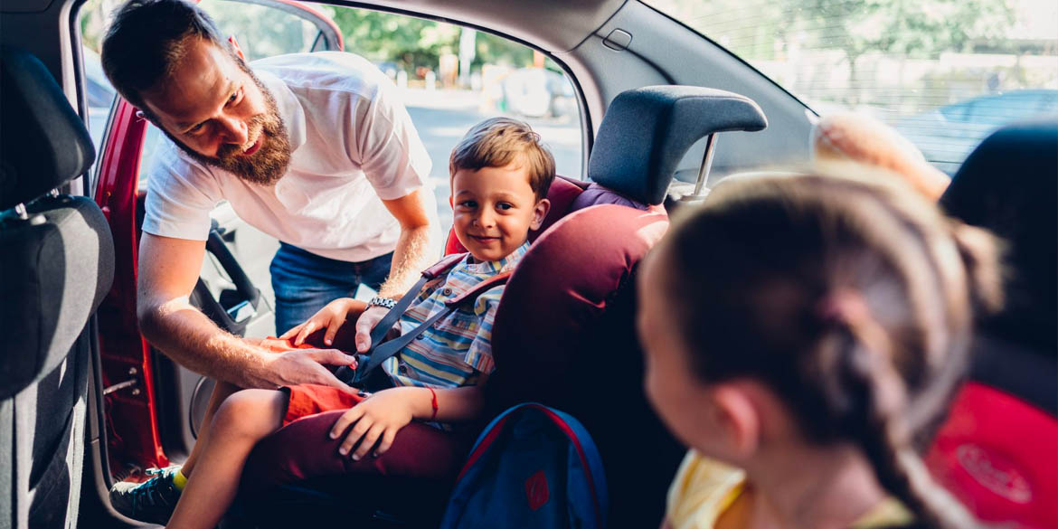 Man in white shirt putting kids into car seats in the back seat with red interior.