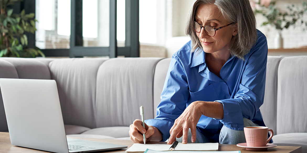 Woman with Gray Hair in Blue Shirt Writing Letter of Instruction with Pen and Laptop on Gray Couch.