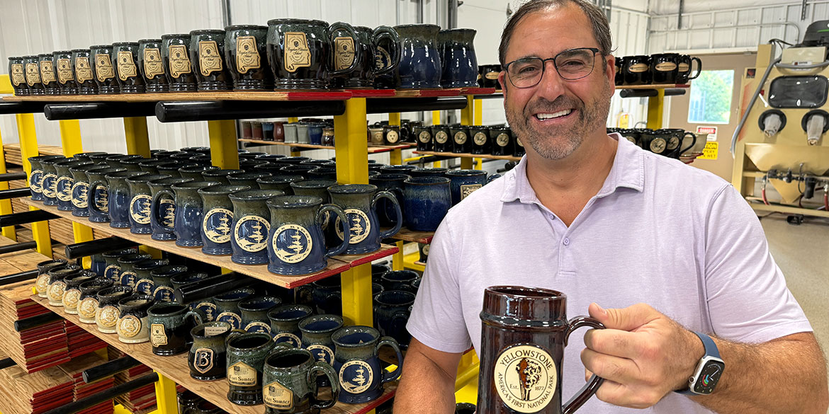Tom Dunsirn holding a mug from Sunset Hill Stoneware with shelves of mugs behind him