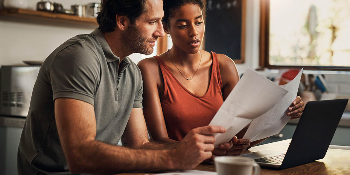 Man in gray shirt with woman in orange shirt in front of laptop comparing papers in kitchen.